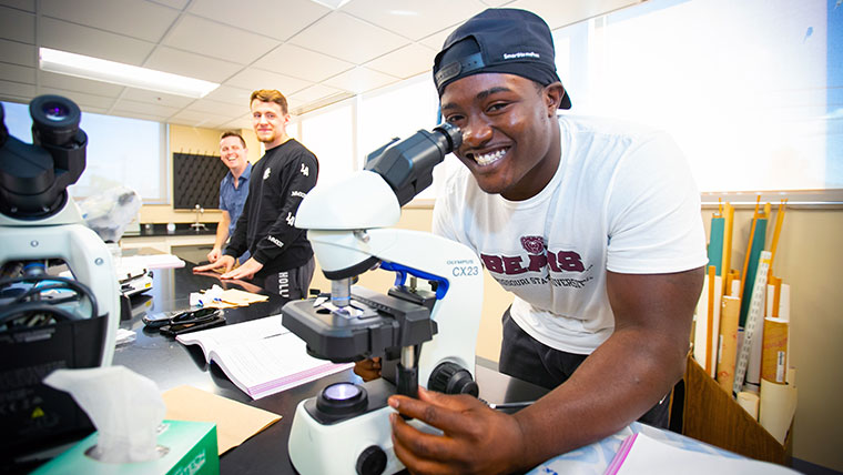A smiling Missouri State student looking through a microscope within a lab. 