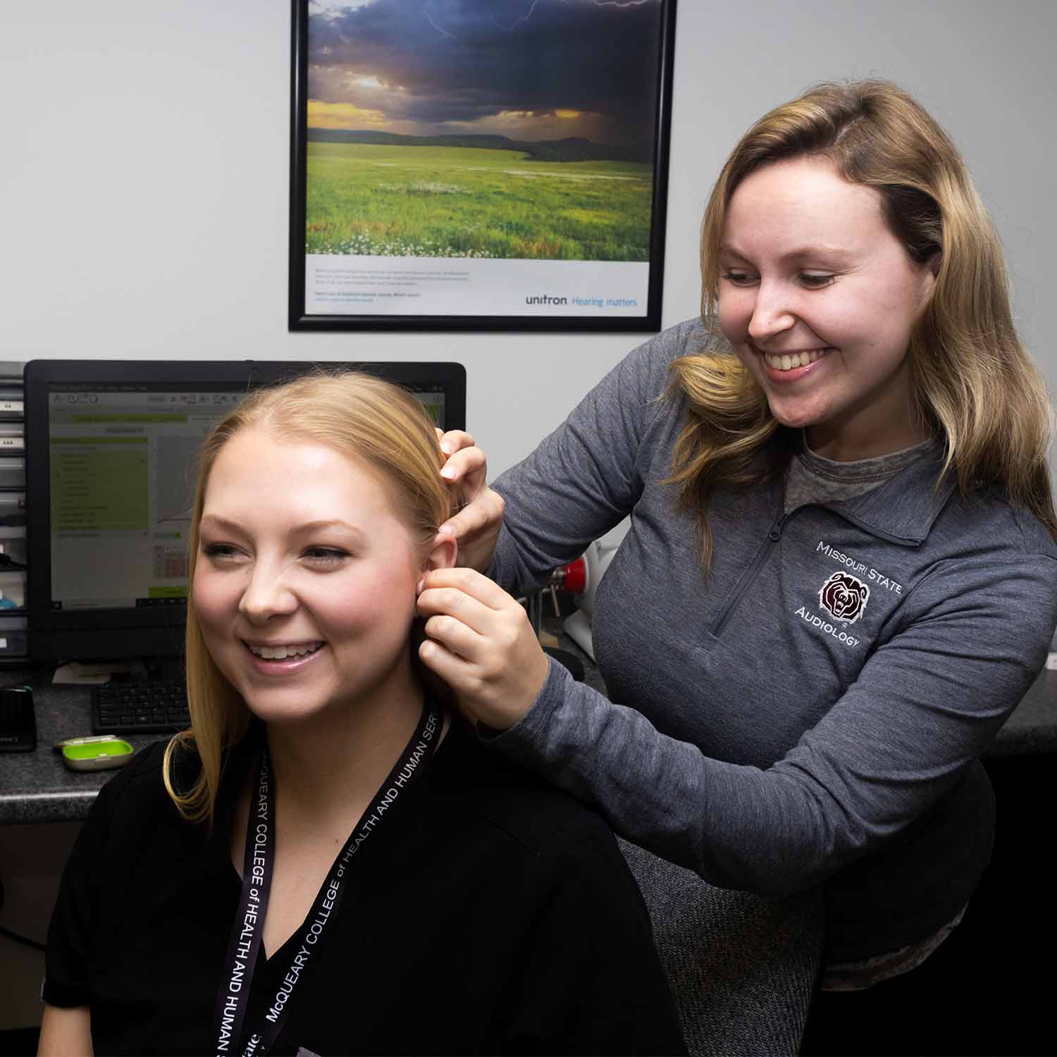 An audiology student connects a hearing device to another student's ear during a lab demonstration.