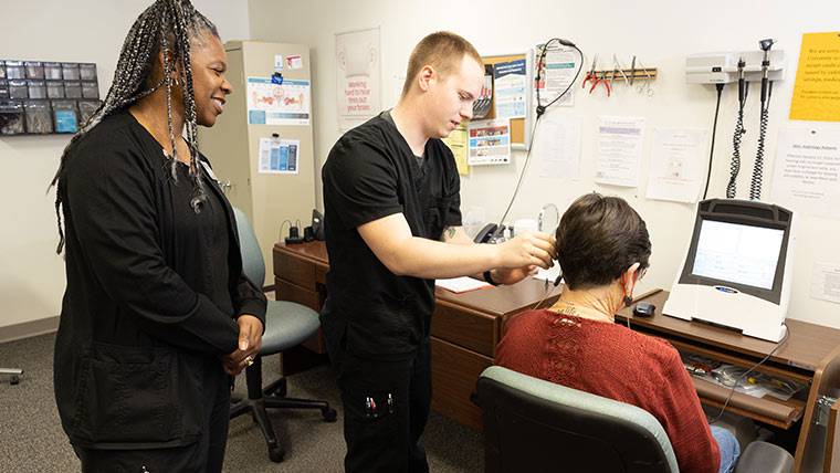 An audiology supervises a student helping a patient put on a hearing device.
