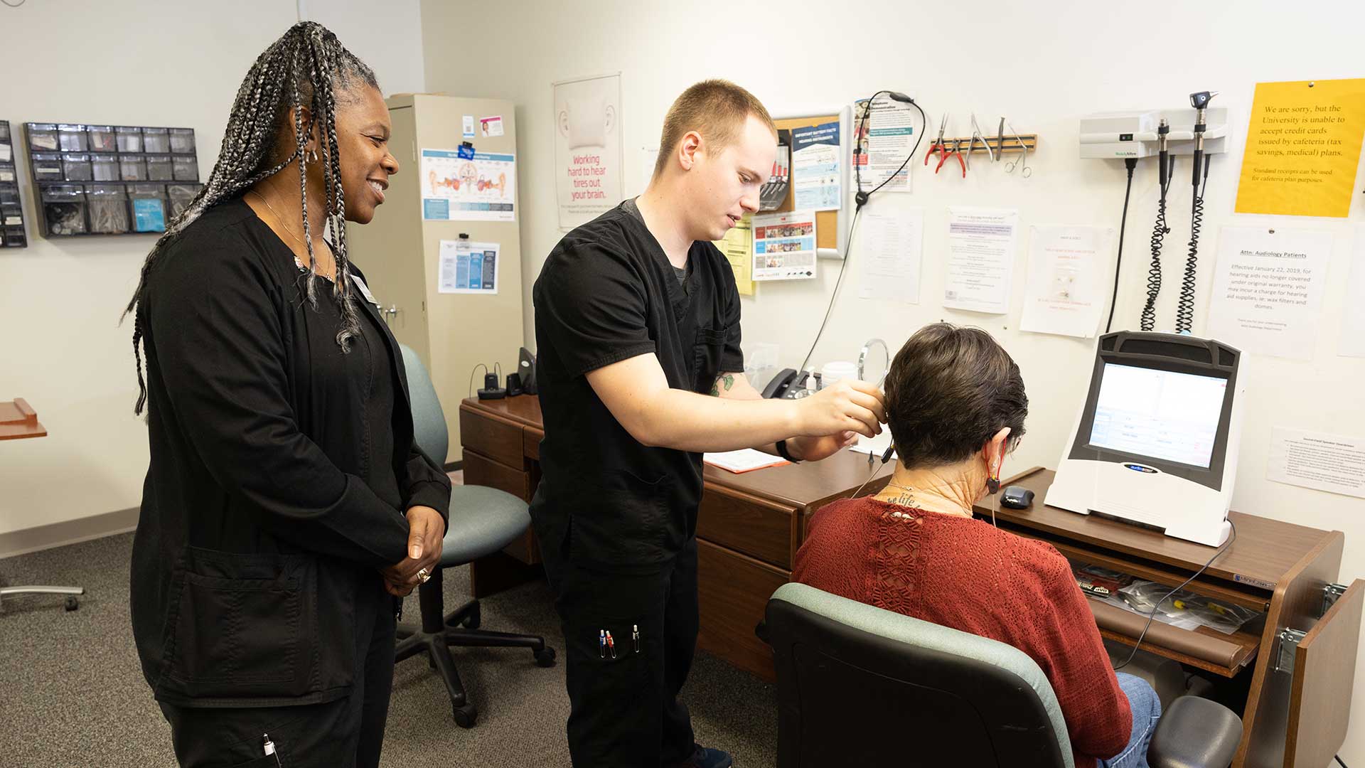 An audiology supervises a student helping a patient put on a hearing device.