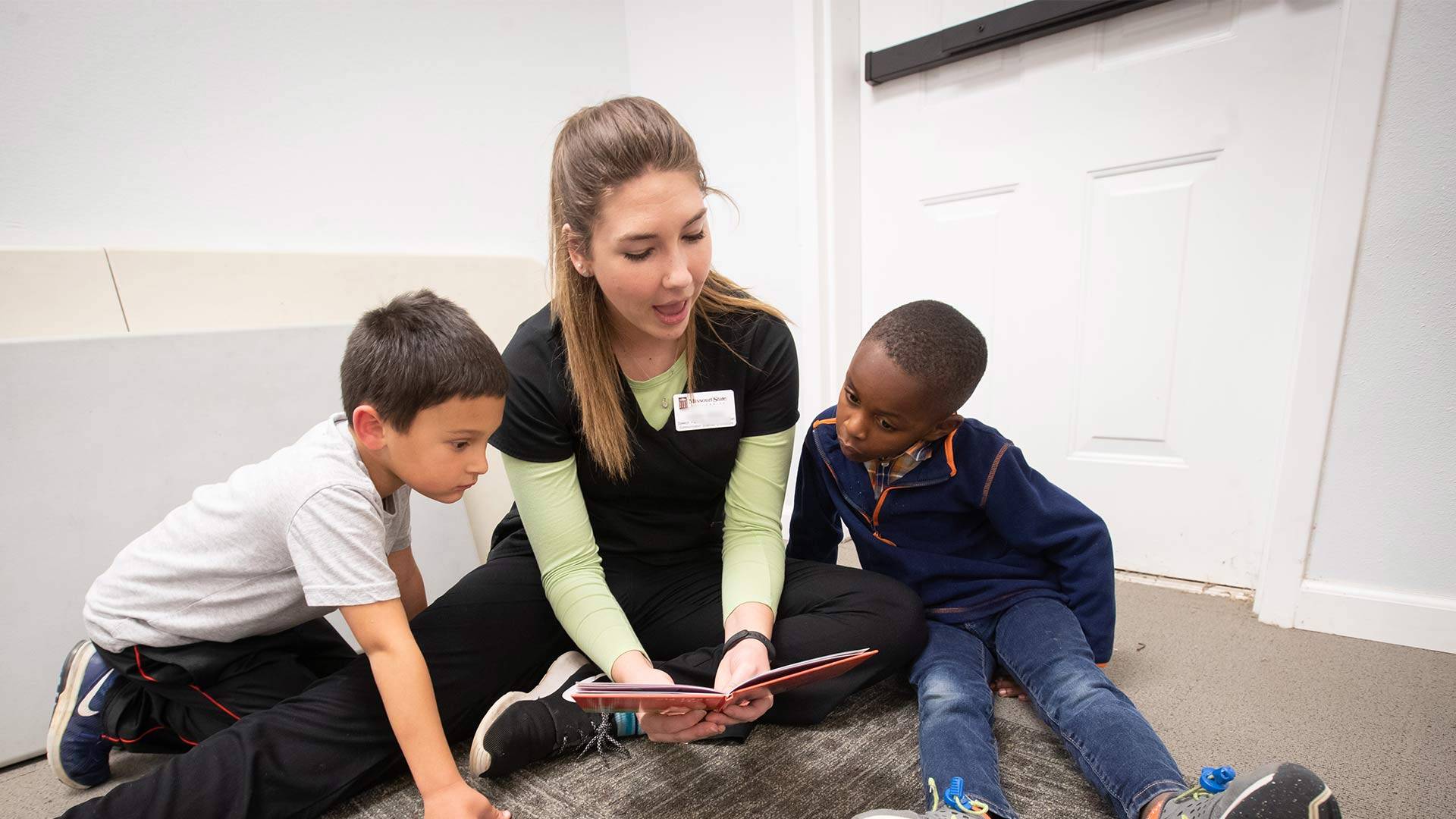 A speech-language pathology student reads a book to two young children at a community event.