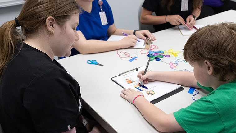 A child draws and colors on a sheet of paper as a student observes.