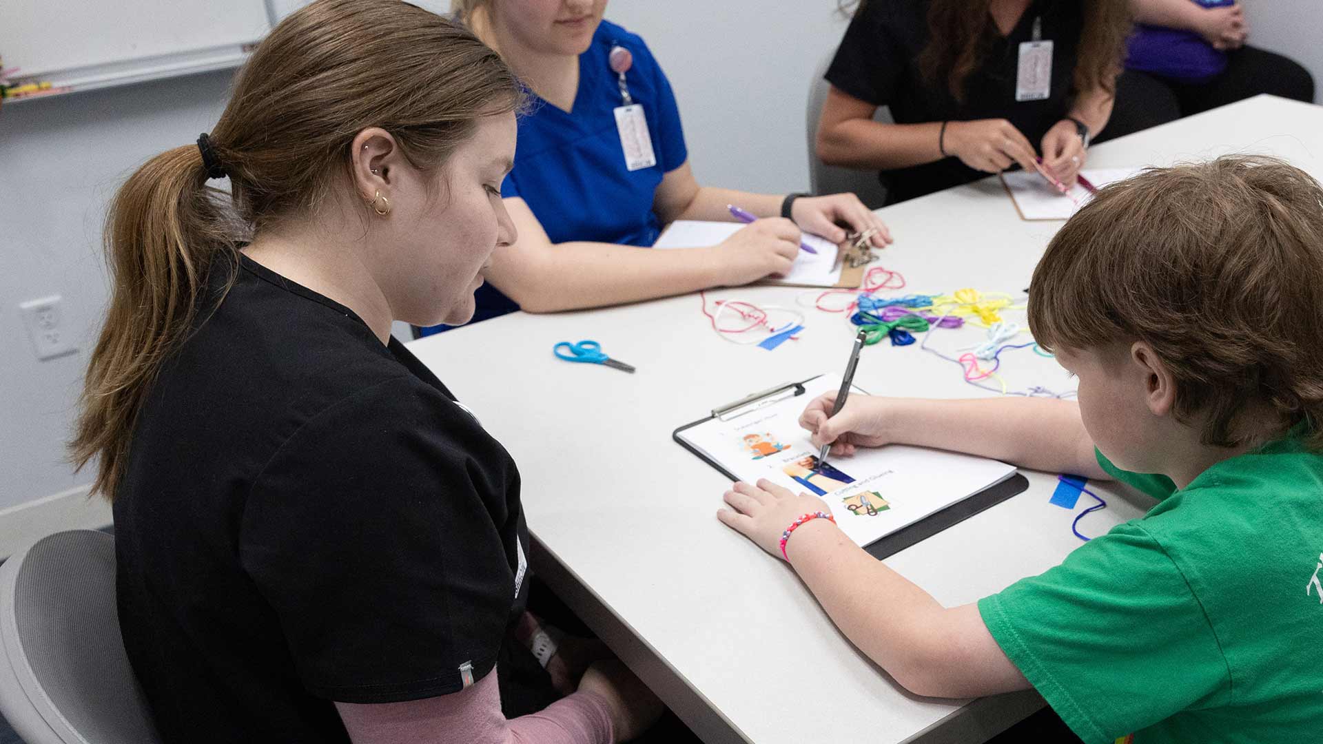A child draws and colors on a sheet of paper as a student observes.